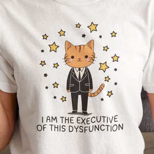 May include: White t-shirt featuring a cartoon cat in a suit surrounded by stars. The cat has orange fur and is wearing a black suit. Text on the shirt reads, "I AM THE EXECUTIVE OF THIS DYSFUNCTION."