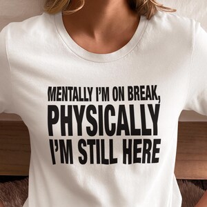 Puede incluir: Camiseta blanca con el texto "MENTALLY I'M ON BREAK, PHYSICALLY I'M STILL HERE" en letras negras en negrita. La camiseta es de cuello redondo. La persona lleva pantalones negros.