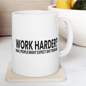 Puede incluir: Taza de cerámica blanca con el texto "WORK HARDER? NAH, PEOPLE MIGHT EXPECT SHIT FROM ME" en negro. La taza está sobre un posavasos beige. La taza tiene un asa curva.