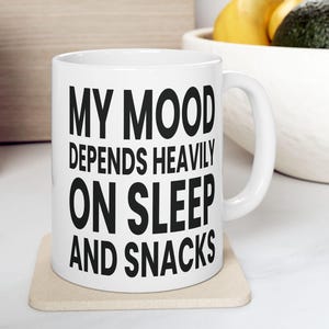 Pode incluir: Caneca de cerâmica branca com o texto "MY MOOD DEPENDS HEAVILY ON SLEEP AND SNACKS" em letras pretas em negrito. A caneca está sobre um descanso de copo bege. O fundo inclui uma tábua de cortar de madeira e uma tigela de frutas.