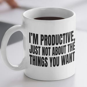Może przedstawiać: Biały ceramiczny kubek wypełniony ciemnym płynem, z czarnym napisem "I'M PRODUCTIVE, JUST NOT ABOUT THE THINGS YOU WANT". Kubek stoi na białym biurku z myszą i notatnikiem.