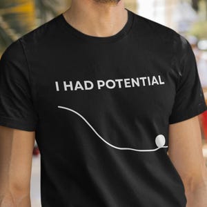 Könnte beinhalten: Schwarzes T-Shirt mit dem weißen Text "I HAD POTENTIAL" über einer gebogenen Linie und einem kleinen weißen Ball. Die Person trägt das T-Shirt im Freien, mit Palmen im Hintergrund.