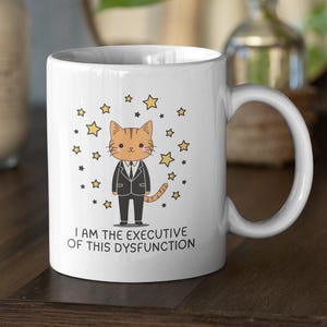 May include: White ceramic mug featuring a cartoon cat in a suit surrounded by stars. The text on the mug reads "I AM THE EXECUTIVE OF THIS DYSFUNCTION". The mug has a curved handle and is set on a dark wooden surface.