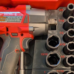 May include: A red and gray Milwaukee impact wrench with a set of black sockets in a red case. The sockets are labeled with sizes like 3/8, 7/16, 1/2, 9/16, 5/8, 15/16, and 1 1/16 inches. The wrench has a warning label.