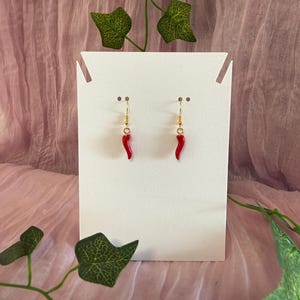 Könnte beinhalten: Ohrringe mit kleinen, leuchtend roten Chili-Pfeffer-Anhängern. Die Ohrringe hängen an goldfarbenen Haken und werden auf einer weißen Karte vor einem weichen rosa Hintergrund mit grünen Efeu-Akzenten präsentiert.