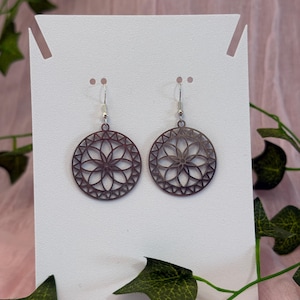 May include: Two dark brown, circular earrings with a floral design are displayed on a white card. The earrings have a filigree pattern and are suspended from silver-colored hooks. Green leaves and vines are in the background.