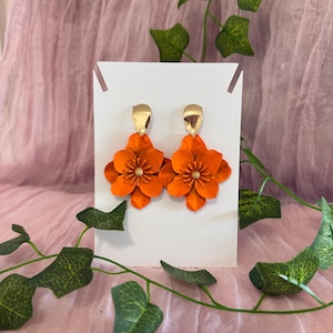 Könnte beinhalten: Ein Paar orangefarbene Blumenohrringe mit goldfarbenen Pfosten. Die Ohrringe werden auf einer weißen Karte vor einem rosa Hintergrund mit grünem Efeu präsentiert.