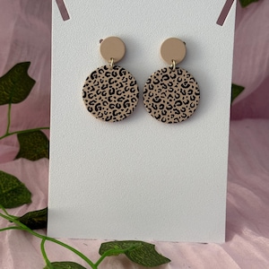 May include: A pair of dangle earrings featuring a small solid beige circle at the top connected to a larger circular pendant below. The larger circles display a black and beige leopard print pattern. The earrings are attached to a white display card.