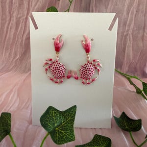 Könnte beinhalten: Ein Paar rosa Krebs-Ohrringe. Jeder Ohrring hat ein Krebsdesign mit einem rosa und weiß gepunkteten Körper, rosa Zangen und einem goldfarbenen Haken. Die Ohrringe werden auf einer weißen Karte vor einem rosa Hintergrund mit grünem Efeu präsentiert.