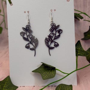 Peut inclure: Une paire de boucles d'oreilles florales gris foncé. Chaque boucle d'oreille présente un motif délicat et stylisé d'une branche fleurie avec de petites fleurs rondes. Les boucles d'oreilles sont suspendues à des crochets d'oreille argentés, présentées sur une carte blanche.