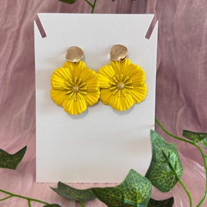 Könnte beinhalten: Ein Paar leuchtend gelbe Blumenohrringe mit goldfarbenen Scheibensteckern. Die Ohrringe werden auf einer weißen Karte präsentiert, mit grünen Blättern und rosa Stoff im Hintergrund. Die Ohrringe haben ein detailliertes Blumendesign.