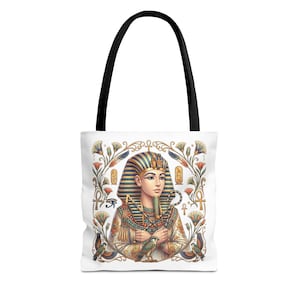 May include: White tote bag featuring a detailed illustration of an Egyptian pharaoh. The pharaoh is adorned with a striped headdress, gold jewelry, and holds a falcon. The bag has black handles and a decorative border with hieroglyphs and flowers.