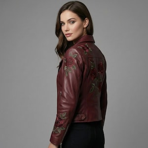 May include: A deep burgundy leather jacket featuring intricate floral embroidery of red roses with green leaves on the back and sleeves. The jacket has a structured collar, a zippered front closure, and a fitted silhouette with long sleeves.