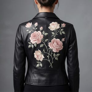 Op de afbeelding: Een zwart leren jasje met een gedetailleerd borduursel van lichtroze rozen en groene bladeren op de achterkant. Het jasje heeft een klassieke kraag, lange mouwen en een getailleerd silhouet met ingewikkeld bloemenstiksel op het achterpand.