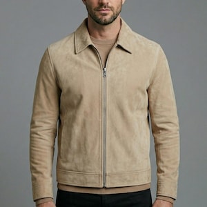 Può includere: Una giacca in camoscio beige con colletto classico, maniche lunghe con polsini a coste e una chiusura centrale con zip in metallo argentato. La giacca è indossata sopra un maglione in maglia beige coordinato con scollo rotondo.