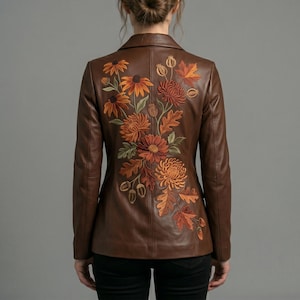 Op de afbeelding: Een bruine leren blazer met een gedetailleerd borduursel op de rug van herfstkleurige bloemen, waaronder oranje en roestkleurige chrysanten, esdoornbladeren en kleine eikels. De jas heeft lange mouwen, een gestructureerde kraag en een getailleerd model.