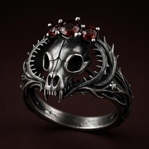 Könnte beinhalten: Ein silberner Ring mit einem Katzenschädel-Design, gekrönt von drei roten Edelsteinen. Der Ring weist detaillierte Dornenranken und scharfe Zähne auf, was ihm einen gotischen oder Fantasy-Look verleiht. Der Ring ist vor einem dunklen Hintergrund platziert.