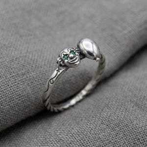 May include: A silver ring featuring a sculpted clown face with small green gemstone eyes and a smooth, rounded balloon shape attached to the side. The band has a twisted, rope-like texture and engraved line details along the outer surface.