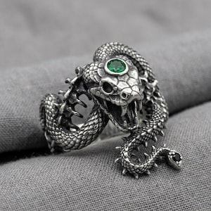 Può includere: Un anello color argento a forma di serpente avvolto con squame testurizzate e punte. La testa del serpente presenta una pietra preziosa verde rotonda e sfaccettata sulla parte superiore, mentre la coda è decorata con una piccola pietra nera.