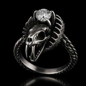 Könnte beinhalten: Silberner Ring mit einem detaillierten Schlangenschädel-Design. Der Schädel hat offene Kiefer mit Zähnen und wird von einem klaren, runden Edelstein gekrönt. Das Ringband hat eine geflochtene Textur.