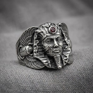 May include: A silver ring featuring a detailed pharaoh head with a small, round, dark red gemstone set in the center of the headdress. The sides of the ring display intricate carvings of winged scarab beetles with textured wings and bodies.