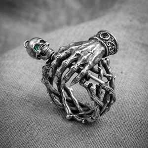 May include: A silver ring featuring a detailed skeletal hand grasping a small key, with a skull at the top set with a vibrant green gemstone. The band is constructed from intertwined, thorny branches, and a dark stone is set into the cuff of the hand.