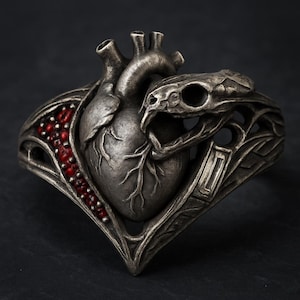 May include: A silver-toned bracelet featuring a detailed anatomical heart design, with a snake head wrapping around it. The bracelet has red accents and intricate metalwork, creating a gothic or fantasy aesthetic.