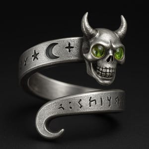 May include: A silver-toned ring featuring a skull with green gemstone eyes and curved horns. The ring has a spiral design with etched symbols, including a crescent moon, star, and cross. The skull has a detailed, grinning expression.