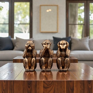 Puede incluir: Tres figuras de monos de color bronce, cada una en una pose diferente: cubriendo los ojos, la boca y las orejas. Las estatuas están sobre una mesa de centro de madera, con un sofá y ventanas en el fondo.