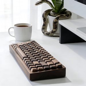 Puede incluir: Un teclado de madera con teclas marrones oscuras y un marco a juego está sobre una superficie blanca. A la izquierda hay una taza de café blanca, y una serpiente está enrollada alrededor de una planta en maceta en el fondo.