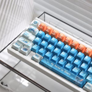 Może przedstawiać: Biała klawiatura mechaniczna z niestandardowymi nakładkami na klawisze. Nakładki na klawisze są niebieskie, pomarańczowe i białe z projektem postaci z kreskówek. Klawiatura ma tekst "My Name is Stitch" na spacji.