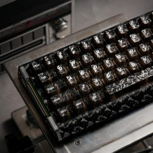 Può includere: Una tastiera meccanica con un motivo mimetico nero e grigio. I copritasti sono trasparenti con lettere bianche. La tastiera ha una cornice metallica ed è posizionata su una superficie metallica. I tasti includono Esc, Tab, Caps, Shift, Ctrl e Win.