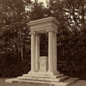 May include: A white marble structure with four columns and a square base. The structure is surrounded by trees.