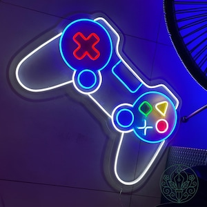 May include: Neon sign in the shape of a video game controller. The controller is outlined in white and blue neon, with red, green, and yellow accents. The background is a dark blue.