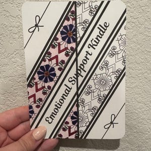 May include: A rectangular card with rounded corners, featuring the text "Emotional Support Kindle." The card has diagonal stripes with floral and geometric patterns in pink, purple, and black. Small bow illustrations are at the top and bottom.
