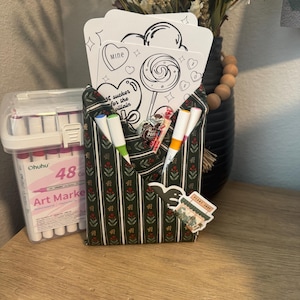 May include: A fabric organizer with a green and red floral pattern, holding white coloring pages with lollipop and heart designs. Markers and a clear case of markers are visible. The text on the coloring pages says "Mine" and "I'm a sucker for the villain".