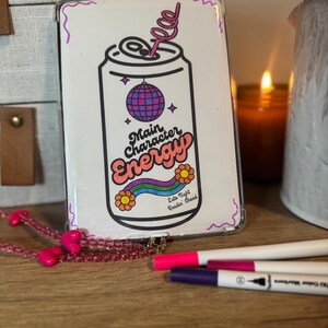 Puede incluir: Una funda transparente para teléfono con un diseño de lata de refresco de dibujos animados con las palabras "Main Character Energy". El diseño incluye una bola de discoteca, un arcoíris y flores. También se ven un collar de cuentas rosas y rotuladores de colores.