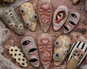 May include: A selection of elaborately decorated artificial nails. The designs include eyeballs, floral patterns, and anatomical features. Some nails are in shades of grey, pink, and white, while others have a black and white striped design.