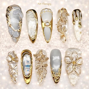 Puede incluir: Una colección de diez uñas artificiales ornamentadas, largas y en forma de almendra. Cada uña presenta un diseño único con detalles dorados, incluyendo patrones florales, adornos de perlas y formas abstractas. El texto "Art Therapy" es visible en la parte superior.