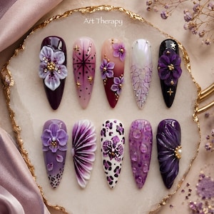 Pode incluir: Um conjunto de dez unhas postiças em formato de amêndoa com designs em roxo, branco e preto. A coleção inclui detalhes florais em 3D, estampa de leopardo, motivos de estrelas e texturas. O texto Art Therapy está impresso no topo central.