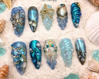May include: A collection of ten long, decorated artificial nails in shades of blue, teal, and white, adorned with pearls, gold accents, and sea-themed designs. The text "Art Therapy" is visible. The nails are arranged on a bed of sand with seashells.