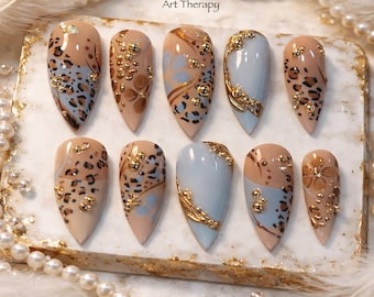 May include: A set of ten artificial nails with various designs. The nails feature a combination of tan, light blue, and gold accents. Some nails have leopard print, while others have floral or abstract patterns. The text "Art Therapy" is visible.