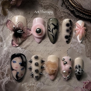 May include: A set of ten press-on nails labeled Art Therapy, featuring diverse designs including a translucent purple flower, a pink button, black botanical art, black polka dots, a portrait of two faces, silver spikes, a peach flower, and silver beads.