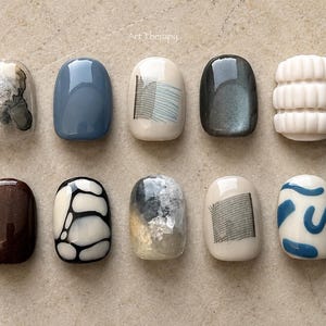 May include: A selection of ten artificial nails with diverse designs. The nails showcase solid colours, abstract patterns, and textured elements. Colours include shades of blue, brown, white, and grey. The text "Art Therapy" is visible in the upper right corner.