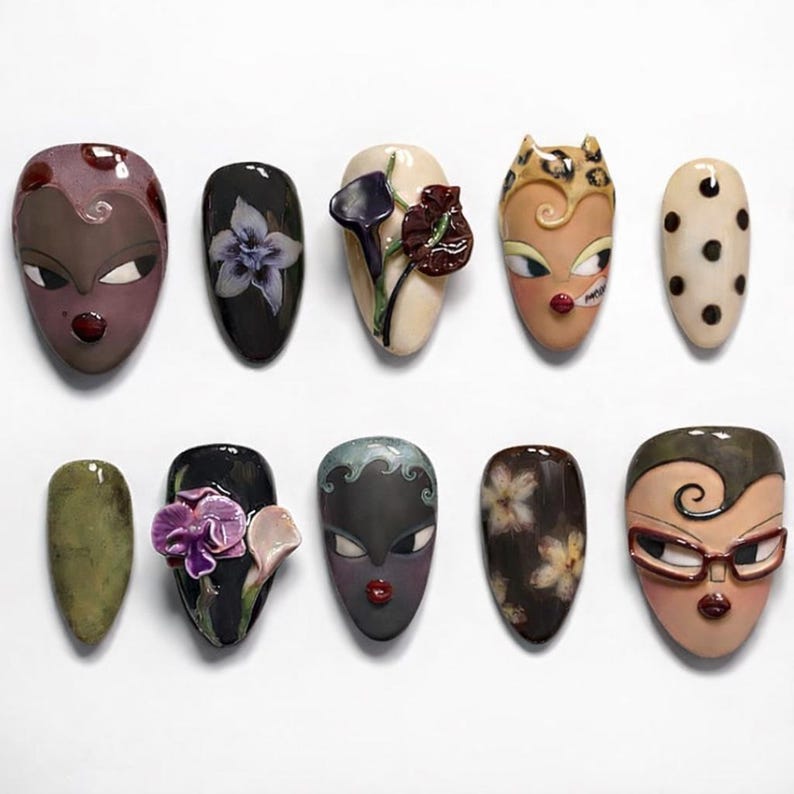 May include: A collection of ten uniquely designed artificial nails. Each nail features a different face design, floral pattern, or abstract art. Colors include shades of brown, black, beige, green, and purple. Some nails have 3D elements.
