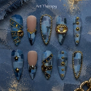 May include: A set of ten stiletto-shaped press-on nails featuring a blue denim patchwork pattern, gold-toned metal studs, star-shaped charms, and buckle accents. The text Art Therapy is visible at the top center of the blue textured background.