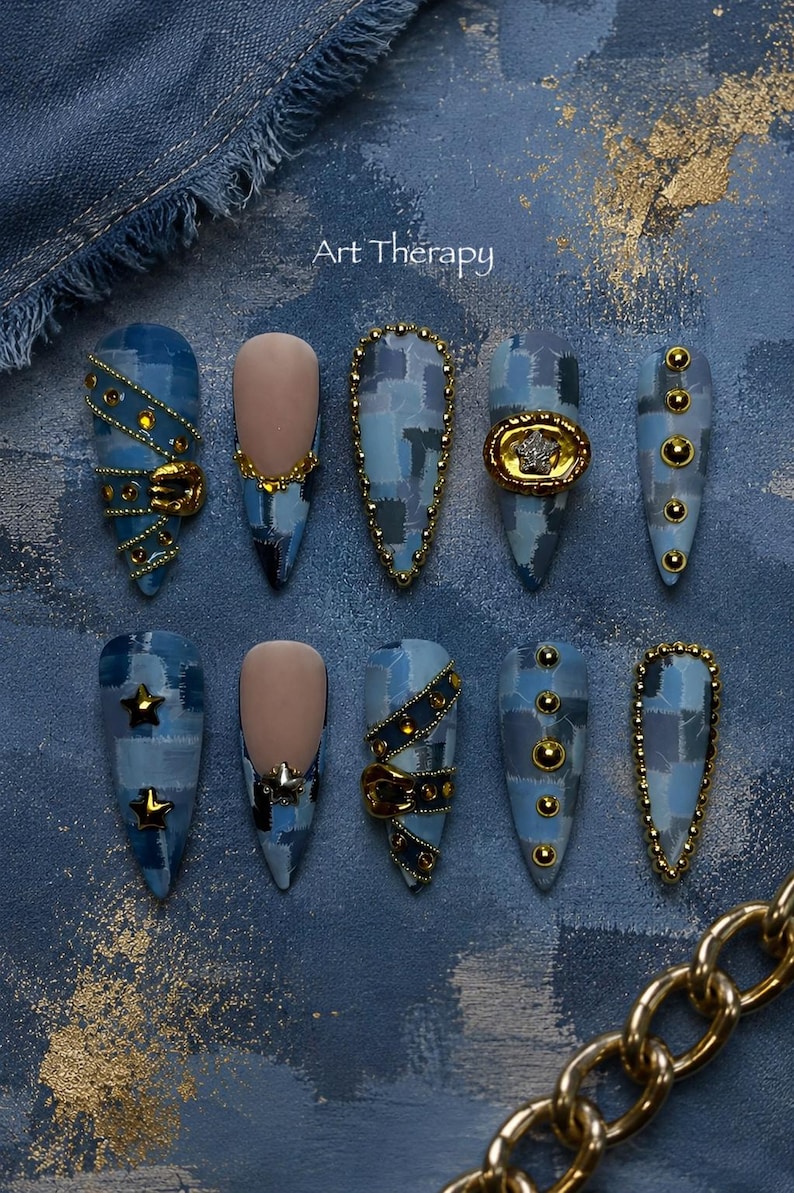 May include: A set of ten stiletto-shaped press-on nails featuring a blue denim patchwork pattern, gold-toned metal studs, star-shaped charms, and buckle accents. The text Art Therapy is visible at the top center of the blue textured background.