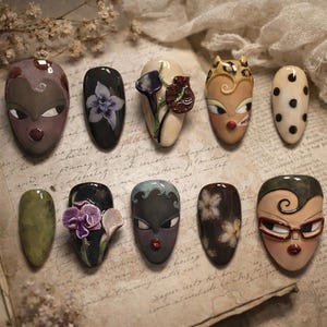 May include: A collection of ten artificial nails with various designs. The nails feature painted faces, floral patterns, and abstract art. Colors include beige, black, brown, and green. Some nails have 3D embellishments.