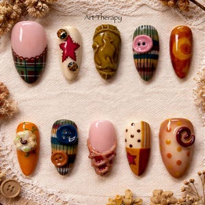 Könnte beinhalten: Eine Sammlung von zehn einzigartig gestalteten Kunstnägeln. Jeder Nagel hat ein anderes Design, einschließlich Muster, Knöpfe und dekorative Elemente. Der Text "Art Therapy" ist oben sichtbar.