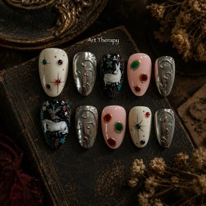 Op de afbeelding: Een set van tien kunstnagels met diverse ontwerpen, waaronder crèmekleurige nagels met rode en groene steraccenten, zilveren metallic nagels met reliëfkrullen, zwarte nagels met witte konijnillustraties en roze nagels met rode en groene ronde edelstenen.